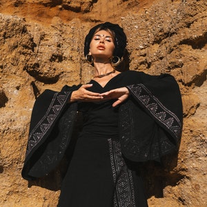 Op de afbeelding: Een zwarte outfit met een top met lange mouwen en een zwierige rok, versierd met ingewikkelde geometrische patronen in een lichtere tint. De outfit wordt gecompleteerd door een zwarte hoofdbekleding, grote gouden oorringen en een delicate ketting. De achtergrond is een getextureerde, aarde-kleurige rotswand.