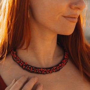 May include: A close-up of a red and black necklace. The necklace features red rectangular beads and small gold-coloured beads, all set on a black woven base. The necklace is around a person's neck.