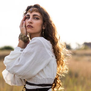 Puede incluir: Una mujer con el pelo largo y rizado lleva una diadema dorada con una piedra azul, una pulsera dorada trenzada y pendientes de aro dorados. Lleva una blusa blanca de manga larga y un cinturón marrón.
