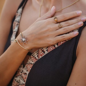 Peut inclure: Un bracelet manchette doré en forme d'éclat de soleil avec une pierre blanche centrale et des bagues assorties. Une bague présente un motif de croissant de lune et d'étoile, l'autre un motif solaire. Les bijoux sont portés avec un haut noir et à motifs.