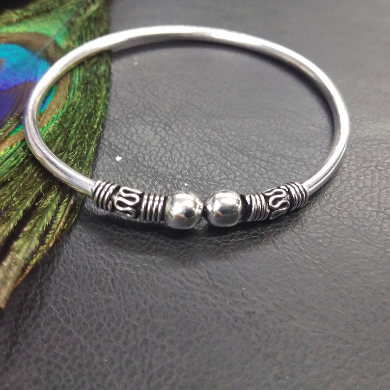 May include: A silver-colored bangle bracelet with decorative detailing. The bracelet features two silver beads and ornate patterns near the center. The design includes a series of lines and a stylized swirl pattern. The bracelet is displayed on a dark surface.