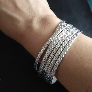 Op de afbeelding: Een stapel zilveren armbanden met een textuur van ruitpatronen. De armbanden worden om een pols gedragen, waardoor hun glanzende, metalen uitstraling wordt benadrukt. Een eenvoudig maar elegant accessoire.