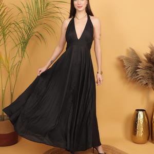 May include: A black halter neck maxi dress with a plunging neckline. The dress has a flowing skirt and is worn with black heels. The background is a solid tan colour with a round jute rug on the floor.