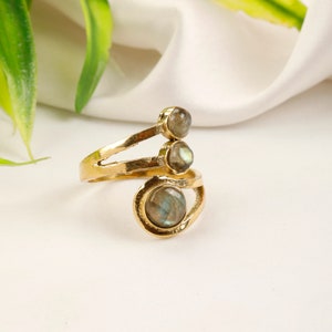 May include: A gold ring featuring three iridescent gemstones. The ring has a distinctive open design, with the gemstones set at different levels. The gemstones display a mix of grey, green, and blue hues. The ring is a piece of jewellery.