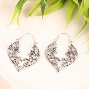 May include: A pair of silver-toned, openwork earrings. Each earring features a heart-shaped design with intricate floral and vine patterns. The earrings have a hook closure and are displayed against a white background with green foliage.