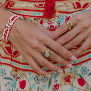 Peut inclure: Un bracelet de perles rouges, blanches et dorées avec un coquillage. Deux bagues dorées sont portées aux doigts. L'arrière-plan présente un tissu à motifs avec des motifs floraux rouges, beiges et bleus.