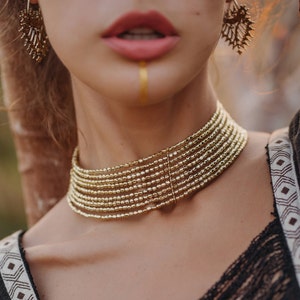 Könnte beinhalten: Ein goldfarbenes Choker-Halsband mit mehreren Strängen. Die Halskette wird am Hals einer Person getragen. Das Bild zeigt auch goldene Ohrringe und eine goldene Linie, die auf das Gesicht der Person gemalt wurde.