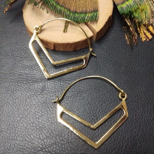 Op de afbeelding: Een paar goudkleurige oorringen. Elke oorring heeft een geometrisch ontwerp met een V-vormig element in een vierkant frame. De oorbellen worden gepresenteerd op een houten oppervlak met decoratieve veren op de achtergrond.