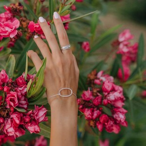 Peut inclure: Un bracelet en argent avec un design ovale est présenté. Une bague en argent assortie est portée au doigt. Les bijoux sont mis en valeur sur un fond de fleurs roses et de feuillage vert. Le bracelet est un bijou délicat.