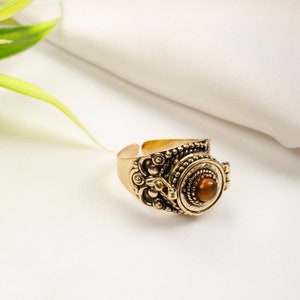 May include: A gold-colored adjustable ring with ornate detailing. The ring features a central, round, brown stone. The design includes intricate patterns and a vintage aesthetic, suitable for jewelry collections.