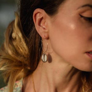 Peut inclure: Boucles d'oreilles créoles dorées avec un pendentif en coquillage blanc et beige. Les boucles d'oreilles sont portées à l'oreille d'une personne à la peau claire et au trait d'eye-liner foncé. Un accessoire simple et élégant.