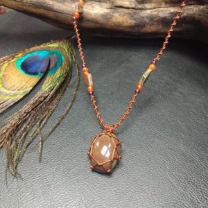 May include: A handcrafted necklace featuring a large, brown gemstone pendant encased in a woven copper wire setting. The necklace has a copper chain with orange and purple beads. A peacock feather and driftwood are in the background.