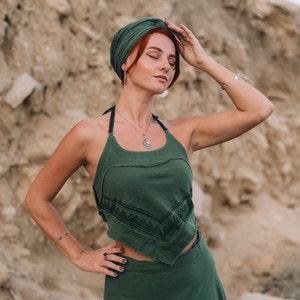 Peut inclure: Une femme porte un haut dos nu vert foncé avec une bordure noire et une jupe assortie. Elle porte également un turban vert, des bijoux en argent et un collier avec un pendentif en forme de croissant de lune. Le fond est de couleur sable neutre.