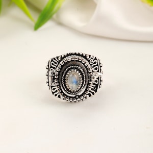 Puede incluir: Anillo plateado con una piedra preciosa ovalada e iridiscente en el centro. El anillo presenta patrones detallados y un diseño texturizado alrededor de la piedra, creando una estética vintage. Se encuentra sobre un fondo blanco.