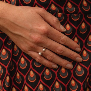 Peut inclure: Une bague argentée ornée de deux sphères. La bague est portée sur une main, avec un tissu à motifs noirs, rouges et oranges en arrière-plan. Le design de la bague est minimaliste et moderne.
