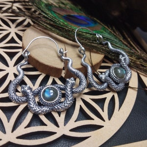 Peut inclure: Boucles d'oreilles serpent argentées avec un motif texturé en écailles. Chaque boucle d'oreille présente une forme de serpent enroulé avec une pierre ronde et irisée au centre. Les boucles d'oreilles sont présentées sur une surface en bois.