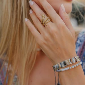 May include: A gold-colored snake ring wraps around a finger, paired with two beaded bracelets. One bracelet features a geometric pattern in gold, black, and white. The other is a light blue bracelet with small white beads.