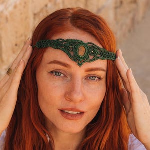 Op de afbeelding: Een donkergroene macramé hoofdband met een ovale steen in het midden en goudkleurige accenten. De hoofdband wordt gedragen door een persoon met rood haar. Het macramé ontwerp is gedetailleerd en creëert een bohemian esthetiek. Een uniek haaraccessoire.