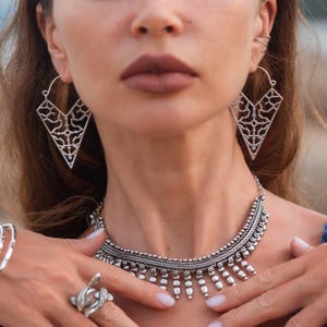 Puede incluir: Juego de joyas plateadas con collar, pendientes y anillo. El collar tiene un diseño detallado con cuentas y detalles colgantes. Los pendientes son grandes formas geométricas caladas. El anillo tiene un diseño único y ondulado.