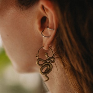 Könnte beinhalten: Nahaufnahme eines goldfarbenen Schlangenohrrings. Der Ohrring ist als Creolen-Design mit einem detaillierten Schlangenanhänger gestaltet. Die Schlange ist gewunden und strukturiert, mit einer Vintage- oder Antik-Ästhetik. Der Ohrring wird an einem Ohr mit einem silbernen Ohrbügel getragen.