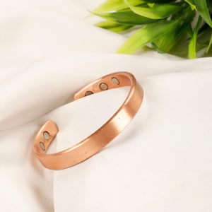 Op de afbeelding: Een koperkleurige manchetarmband met drie kleine, zilverkleurige magneten. De armband heeft een eenvoudig, plat ontwerp en is aan de uiteinden open. De armband wordt gepresenteerd op een wit oppervlak met groen gebladerte op de achtergrond.