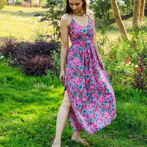 May include: A pink floral print maxi dress with a V-neck and spaghetti straps. The dress has a high slit and is worn with brown sandals. The background is a green garden.