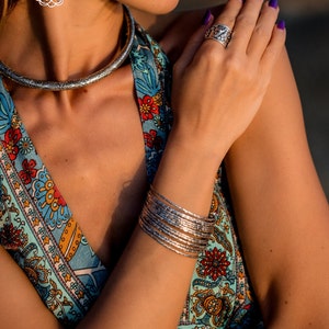 May include: A silver-toned jewelry set featuring a patterned choker necklace, a ring, and multiple bangles. The jewelry is worn with a colorful floral print top. The ring and necklace have detailed designs.