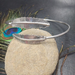 May include: A silver cuff bracelet with two detailed feather motifs. The open design allows for easy wear. The bracelet is displayed on a stone. The feathers are intricately designed, adding a touch of nature-inspired elegance to the jewellery.