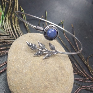 Puede incluir: Un brazalete plateado con una piedra preciosa azul y un diseño de hojas. El brazalete presenta una piedra azul redonda engastada y una rama de hojas decorativa. El brazalete está sobre una superficie de piedra.
