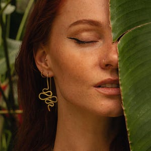 Puede incluir: Pendientes dorados con forma de serpiente. Los pendientes son largos y colgantes, con un diseño de serpiente estilizado. El fondo es una mezcla de follaje verde y un rostro, con un ojo cerrado y un piercing en la nariz.