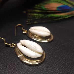 Puede incluir: Un par de pendientes dorados con conchas de cauri blancas. Las conchas están engastadas en marcos ovalados de color latón con cierre de gancho. Los pendientes se exhiben sobre una superficie oscura, con una pluma de pavo real en el fondo.