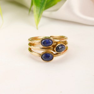 Puede incluir: Anillos dorados con tres bandas, cada uno con una piedra preciosa ovalada de color azul oscuro. Los anillos están apilados, creando un efecto de capas. Las piedras preciosas tienen un acabado pulido y las bandas doradas son lisas y brillantes. Joyas.