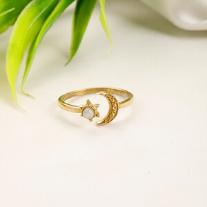 May include: A gold-coloured ring with a crescent moon and star design. The star holds a small, round, white gemstone. The ring is set against a white background with green foliage in the upper left corner.