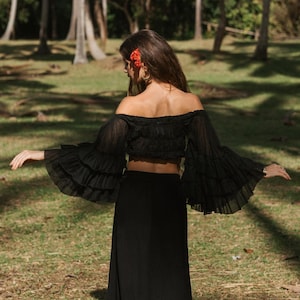 Könnte beinhalten: Schwarzes schulterfreies Crop-Top mit Rüschenärmeln und einem passenden schwarzen Rock. Das Oberteil hat ein gerafftes Oberteil. Das Outfit wird im Freien in einer parkähnlichen Umgebung mit Bäumen und Gras getragen.