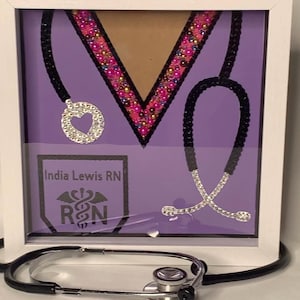 May include: A framed artwork featuring a purple background with a decorative stethoscope design. The design includes a V-shaped element with pink and gold embellishments, and the text "India Lewis RN" and "R&N". A silver stethoscope rests in front.