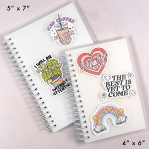 May include: Two white spiral notebooks with various stickers. Stickers include: "Iced Coffee All Year Long", a plant with a rainbow pot, a heart with "Self Love Club", "The Best Is Yet To Come", and a rainbow with books. Notebooks measure 12.7cm x 17.8cm and 10.2cm x 15.2cm.