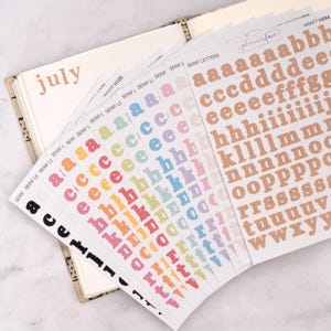 May include: An open planner showing the month of July, with several sticker sheets fanned out around it. The sticker sheets contain serif letters in a variety of colours, including pastels and a kraft paper shade. The stickers are for planners and journals.
