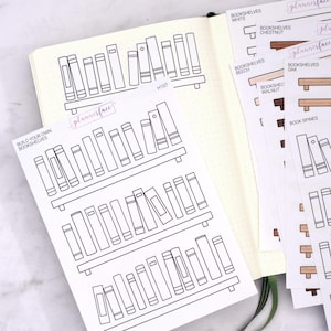 Könnte beinhalten: Eine Planerseite mit einem weißen Notizbuch und Aufkleberbögen. Die Aufkleber zeigen Bücherregal-Designs und den Text "BUILD YOUR OWN BOOKSHELVES" und "plannerface". Zusätzliche Blätter zeigen Bücherregal-Farboptionen und Buchrücken.