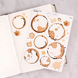 May include: A sheet of coffee stain stickers featuring brown ring and splatter designs. The sheet is labelled "COFFEE STAINS washi paper" and "plannerface". The stickers are next to an open notebook with dotted pages.