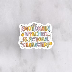 Puede incluir: Una pegatina blanca con el texto "Emotionally Attached to Fictional Characters" en letras coloridas. La pegatina tiene un contorno ondulado, pequeñas flores y caras sonrientes. El fondo es un mármol gris claro.