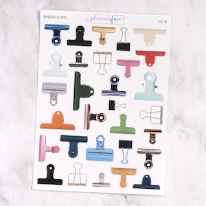 Puede incluir: Una hoja de pegatinas con clips de encuadernación de varios colores, incluyendo azul, negro, dorado, rosa y verde. La hoja tiene el texto "BINDER CLIPS" y "plannerface" impresos.