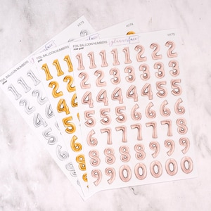 May include: Three sheets of foil balloon number stickers in silver, gold, and rose gold. The stickers feature numbers 0-9 in a balloon style. The text "FOIL BALLOON NUMBERS" and "plannerface" are printed on the sheets.