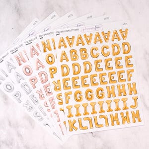 May include: Sheets of foil balloon letter stickers in silver, rose gold, and gold. The gold letters spell out the alphabet. The stickers are labelled "foil balloon letters" and "plannerface".