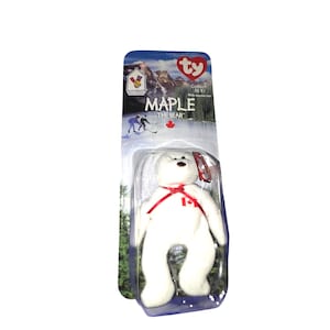 May include: A white plush bear toy named "Maple" with a red ribbon and the Canadian flag on its chest. The toy is in a clear plastic package with a mountain and hockey scene on the backing card. The card reads "Maple The Bear".