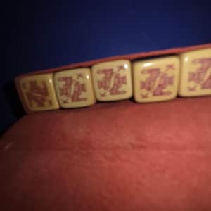 May include: A close-up of a bracelet featuring cream-colored square beads with a red design. The bracelet is resting on a red surface, with a blue background.