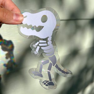 May include: A holographic sticker with a white dinosaur skeleton design. The sticker has a clear, iridescent border and a detailed illustration of the dinosaur's bones. The design is a stylized representation of a Tyrannosaurus Rex.