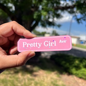 May include: A rectangular pink sticker with a white border and white text that reads Pretty Girl Ave. The sticker features a subtle holographic glitter finish with small star and dot accents scattered across the surface of the pink background.