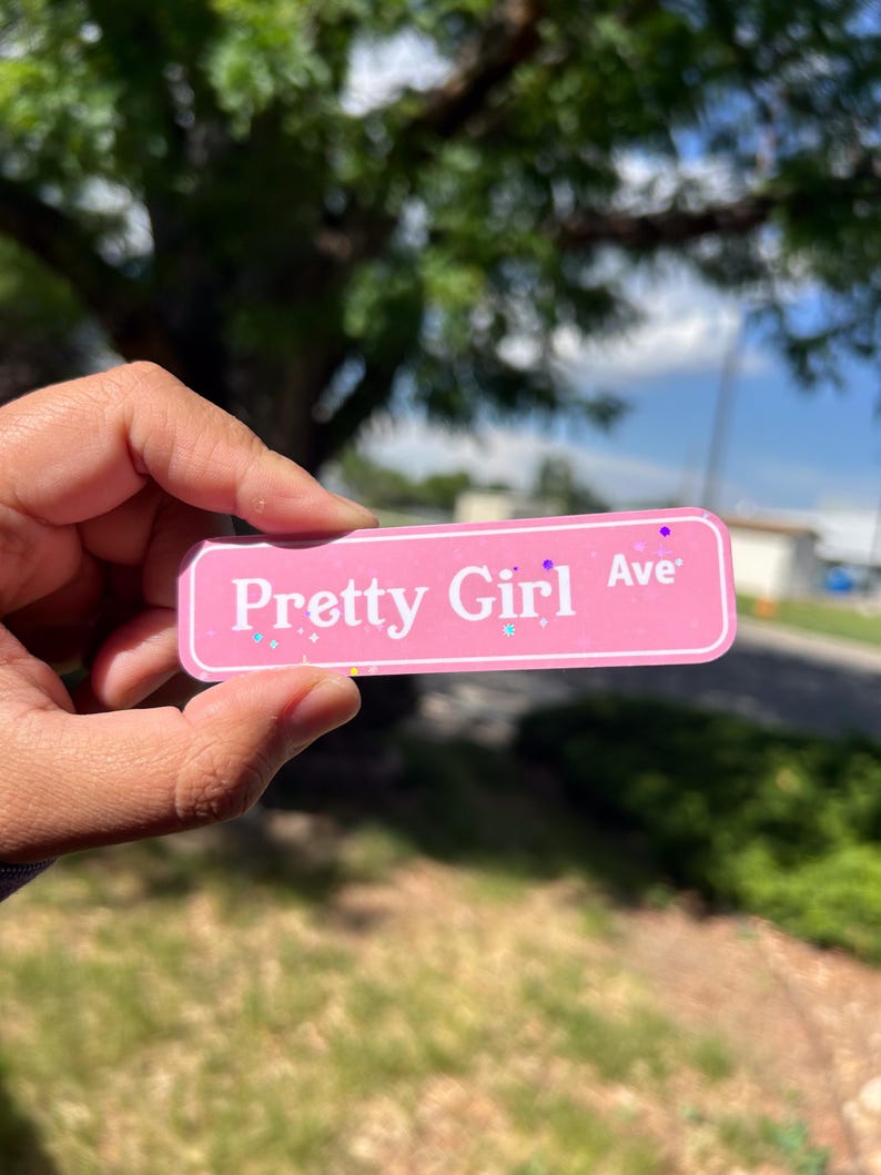May include: A rectangular pink sticker with a white border and white text that reads Pretty Girl Ave. The sticker features a subtle holographic glitter finish with small star and dot accents scattered across the surface of the pink background.