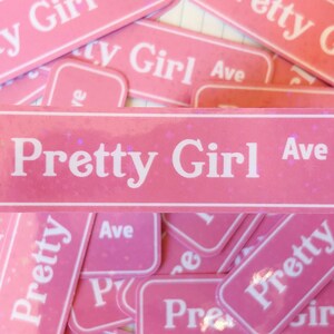 May include: A rectangular pink sticker featuring a white border and the white text Pretty Girl Ave. The sticker has a subtle holographic glitter finish that catches the light, creating a sparkling effect across the surface of the pink background.