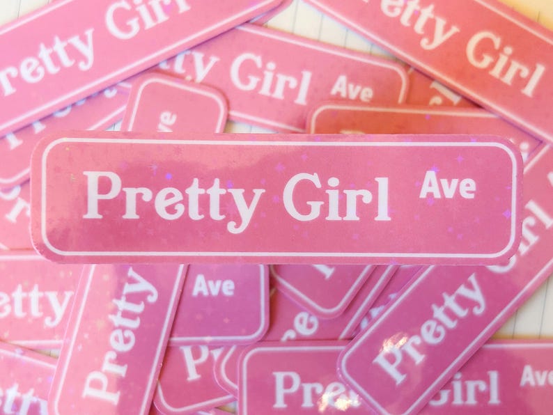 May include: A rectangular pink sticker featuring a white border and the white text Pretty Girl Ave. The sticker has a subtle holographic glitter finish that catches the light, creating a sparkling effect across the surface of the pink background.